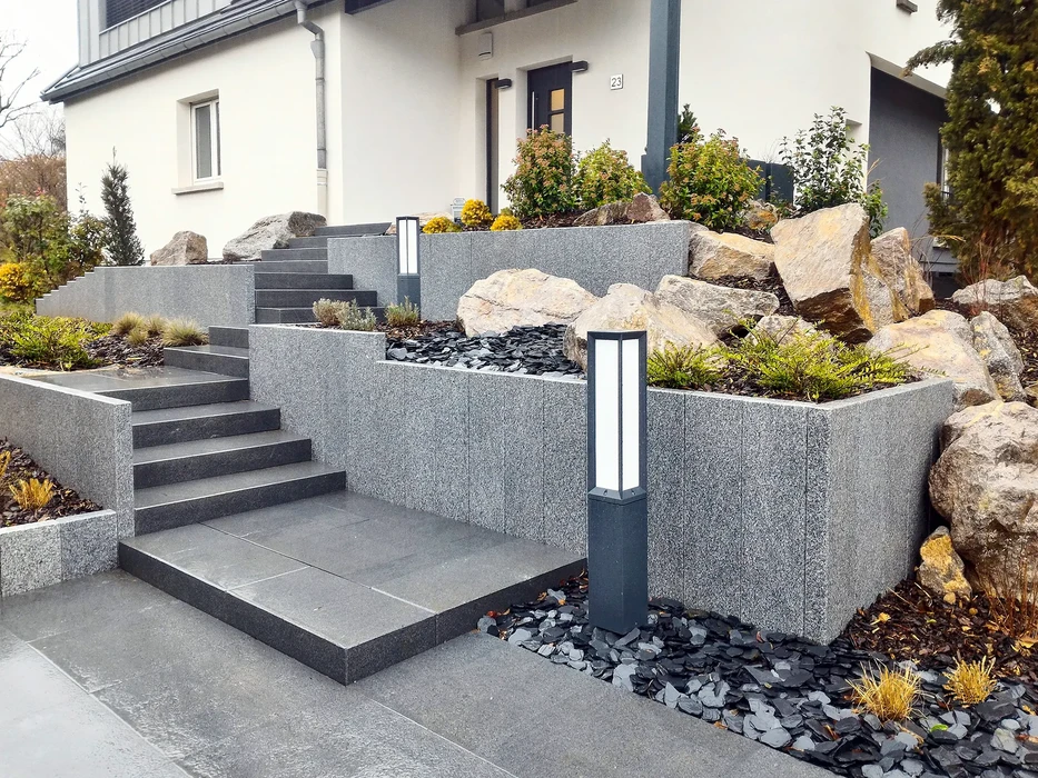 Paysage montagnard avec vallée verdoyante, rivière serpentant au premier plan, cascade en arrière-plan, sommets rocheux enneigés, végétation alpine luxuriante et fleurs roses sauvages